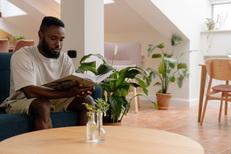 A man relaxes on a couch reading a book in a plant-filled living room.