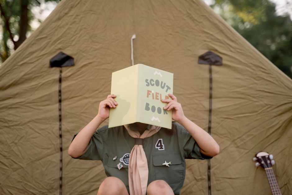 A scout child reads a field book outside a tent, embracing outdoor adventure.